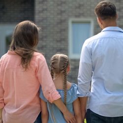 A family of three standing in front of a home looking at it.
